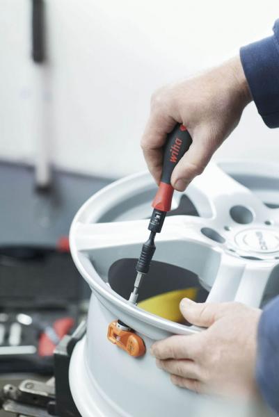 A hand holds a screwdriver tightening a screw on a silver rim. In the background, tools and a bright environment are faintly visible.