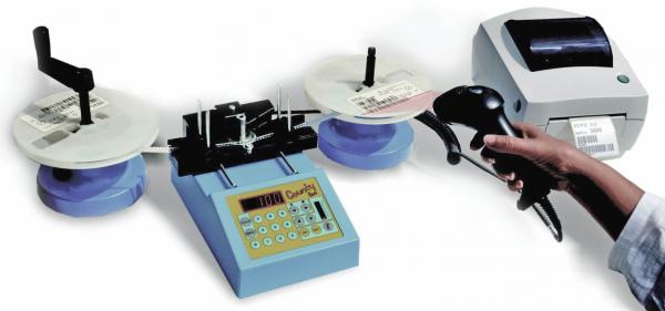The image shows a device setup for labeling. In the center is a blue device with buttons, with rolls of labels and a printer on the left and right. A hand is holding a scanner.