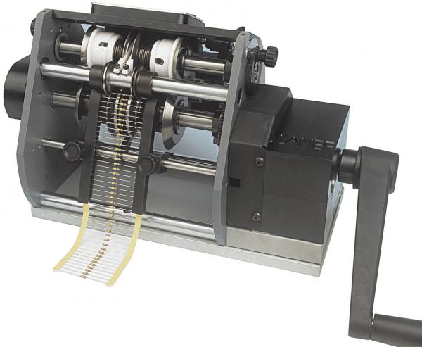 Das Bild zeigt eine mechanische Maschine mit sichtbaren Zahnrädern und Walzen. Eine Handkurbel befindet sich auf der rechten Seite, und ein langer, dünner Streifen Papier zieht sich von der Maschine.