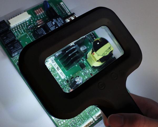 The image shows a hand holding a magnifying glass over a green circuit board. The magnifying glass illuminates an area with various electrical components, including a black element and a yellow one.