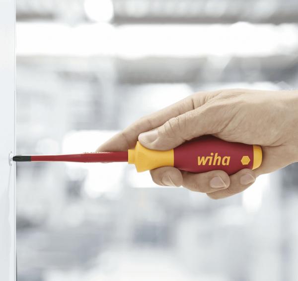 The image shows a hand holding a red and yellow screwdriver. The tip of the screwdriver is black and is being inserted into a screw. The background is blurry and bright.