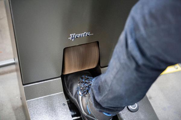 The image shows a foot pressing on a foot pedal. The pedal is attached to a gray, metallic machine that features a logo. In the background, blurred elements are visible.