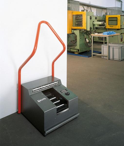 Das Bild zeigt eine graue Maschinenbox mit einem großen roten Griff in Form einer geschwungenen Linie. Im Hintergrund stehen weitere Maschinen in einer Industriehalle.