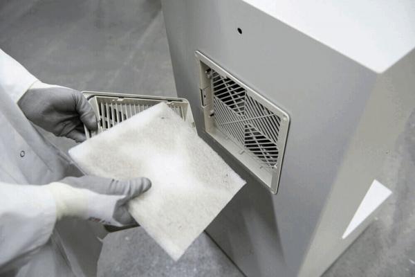 A person in a white coat is holding a dirty air filter with both hands. The filter is rectangular and has a fluffy, gray surface. In the background, there is a device with a ventilation slot.