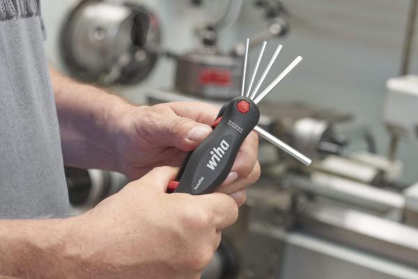 The image shows a person holding a compact toolset in their hand. The handle is black with red accents. Several metal rods protrude from the top end, resembling hex keys. In the background, machines and a workshop can be seen.
