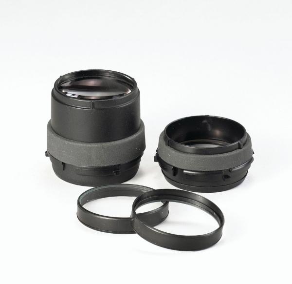 The image shows two black camera lenses with gray rubber coating. Next to them, there are two black filter rings. The objects are arranged on a light surface.
