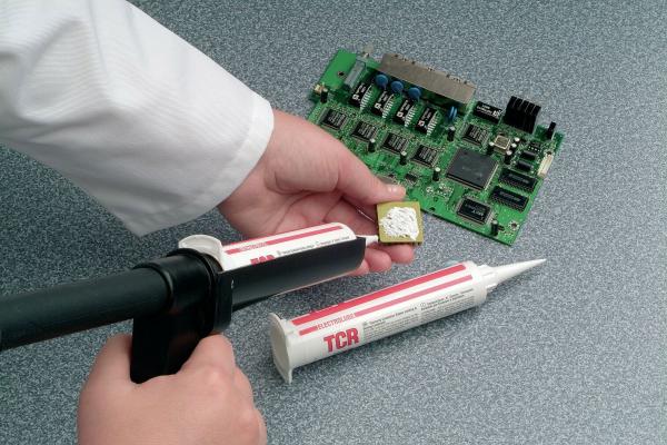 Eine Hand hält eine Leiterplatte mit verschiedenen Bauteilen. In der anderen Hand wird eine Düse verwendet, um etwas Kleber aufzutragen. Zusätzlich liegt eine Tube Kleber auf der Oberfläche.