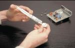 Preview: A person's hands hold a small plastic body while they align a syringe with a silvery substance on it. In the background, there is an electronic component with many small parts.