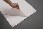 Preview: Auf dem Bild sieht man eine Hand, die eine Ecke eines weißen Blattes anhebt. Darunter ist eine glatte, transparente Folie sichtbar. Der Hintergrund ist grau.