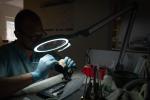 Preview: A man with glasses and rubber gloves is working under a bright magnifying lamp. He is holding a small object in his hand and focusing on his work in a dimly lit room.