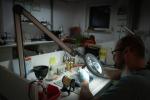 Preview: A man works intently at a table under a large, illuminated magnifying glass. Around him are tools and materials that create a workshop atmosphere.