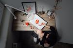 Preview: The image shows a table with art supplies. A person is sitting there, painting with reddish colors. Around the table, there are several objects, including a frame and a plant.