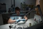 Preview: A man with glasses and gloves is sitting at a table, working under a small, bright lamp. He is focused on an object in his hand, surrounded by tools and materials.
