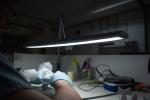 Preview: In the picture, you see a worktable with a bright lamp. A hand wearing blue gloves is holding something small. Various utensils and materials are scattered around the table.