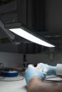 Preview: A table with a bright, swiveling lamp. A hand in blue gloves holds a small object. In the background, tools and equipment are out of focus.