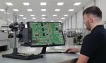 Preview: Ein Mann sitzt an einem Tisch und betrachtet auf einem Monitor eine grüne Leiterplatte mit Bauteilen. Der Raum ist hell und modern, mit weiteren Arbeitsplätzen im Hintergrund.