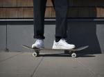 Preview: Auf dem Bild steht eine Person auf einem Skateboard. Sie trägt weiße Sneakers und dunkle, weite Hose. Der Hintergrund besteht aus Holzpaneelen und einer grauen Wand.