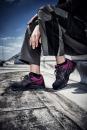Preview: Ein Paar sportliche Schuhe in Schwarz und Pink steht im Fokus. Eine Person sitzt mit einer Hand auf dem Oberschenkel auf einem grauen Untergrund. Sie trägt lockere, graue Hosen. Der Himmel ist blau mit einigen Wolken.