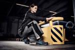 Preview: A sportily dressed woman is kneeling at a yellow device in an industrial hall. She is wearing gray work clothes and looking forward intently. The environment is modern and functional.