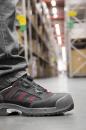 Preview: The image shows a foot wearing a black safety shoe with red accents. In the background, shelves with boxes and a wide warehouse can be seen.