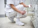 Preview: Eine Person in weißer Kleidung und weißen Schuhen sitzt auf dem Boden und greift in ein Regal, um etwas herauszuholen. Der Boden ist hell und die Umgebung wirkt sauber und ordentlich.
