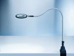 Preview: The image displays a flexible magnifying glass with a round lens at the end of a curved arm. The arm is mounted on a sturdy, rectangular base that supports the lens. The background is bright and monochromatic.