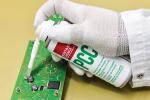 Preview: A hand in a white glove holds a spray can over a green circuit board. The can has red and green lettering. Liquid is being applied to the board.