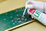 Preview: The image shows a green circuit board on which a person wearing a glove is applying spray. The spray container is labeled and has a white and red color.