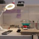 Preview: A workspace with a light magnifier, a digital display, tools in a container, a bowl with green contents, and notes on the wall. Everything neatly arranged.