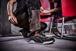 Preview: Ein Mann kniet auf dem Boden in Arbeitskleidung mit dreckigen Hosen und Sicherheitsschuhen. Er greift nach einem Kabel, das zu einem roten Stuhl führt, vor einer rauen Wand.