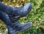 Preview: Das Bild zeigt ein Paar schwarze Wanderschuhe mit einem sportlichen Design. Eine Person trägt schwarze Socken, und im Hintergrund sind grüne, verschwommene Pflanzen zu sehen.