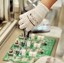 Preview: A hand in a white glove holds a pair of pliers and works on a green circuit board with various components on it. In the background, an industrial environment is visible.