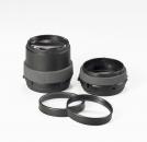 Preview: The image shows two black camera lenses and two black rings. The lenses have a round shape and are partially surrounded by a gray material. They are placed on a white surface.