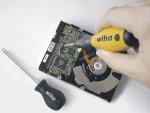 Preview: The image shows a hand pointing a screwdriver with a yellow handle at a hard drive. Next to the hard drive lies another small screwdriver. The surroundings are simple and bright.