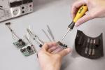 Preview: A hand holds a screwdriver over an electronic circuit board. Next to it, several cables and more circuit boards are lying around. In the background, a measuring device and a tool holder can be seen.