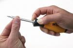 Preview: In the picture, there is a hand holding a screwdriver with a yellow handle. The tip of the screwdriver is pointing upwards, while a second hand carefully holds it steady.