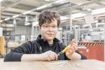 Preview: A teenager with glasses smiles at the table, holding a tool in his hand. Several drawing tools are lying in front of him. In the background, a workshop can be seen.