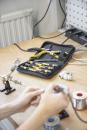 Preview: On a table, there are tools in a black case with yellow handles. One hand is holding a soldering iron, while the other works on a wire. In the background, cables and a holder are visible.
