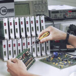 Preview: A hand holds a screwdriver and works on a circuit board. In the background are several devices with red displays, representing test and measurement instruments.