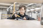 Preview: A boy with short, brown hair and glasses is sitting at a table in a workshop. He is holding a tool in his hand and looking intently at a gray material sample.
