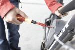 Preview: A hand holds a screwdriver with a red and yellow handle. The screwdriver is pointed at a screw on a black bicycle. The background is bright and blurry.