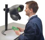 Preview: A man in a blue work coat examines a green circuit board through a large, black magnifying device. The table is plain, with the focus on the work.