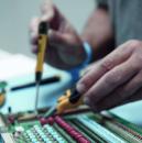 Preview: A person is working on a green circuit board. They are holding a soldering iron in their right hand and using tweezers in their left hand. The board has colorful components.