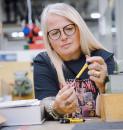 Preview: A woman with long, blonde hair and glasses is sitting at a table in a workshop. She is holding a yellow pen and looking at it intently. In the background, technical equipment is visible.