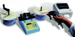 Preview: The image shows an electronic measuring station with a display, connected to two plates and a printer. A hand is holding a barcode scanner.